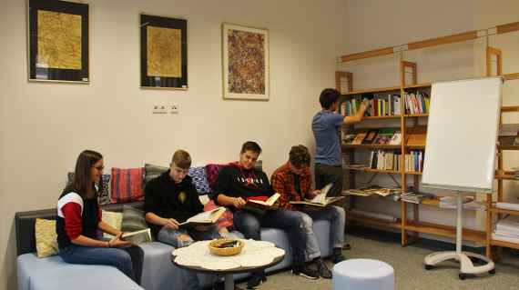 Vier Schülerinnen und Schüler sitzen auf einem blauen Sofa und lesen. Ein Schüler sucht ein Buch bei einem Regal.