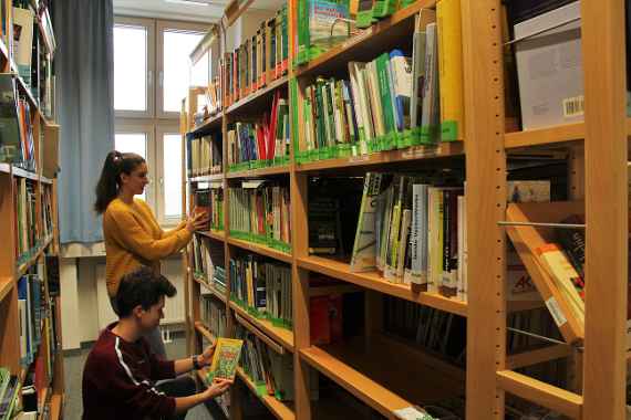 Eine Schülerin und ein Schüler suchen in einem Buchregal ein Buch.