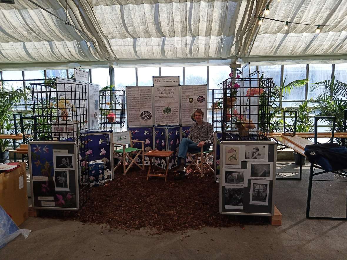 zu sehen ist ein Ausstellungsstand, geschmückt mit Orchideen und verschiedenen Plaktaten. Ein junger Mann sitzt in einem Sessel.