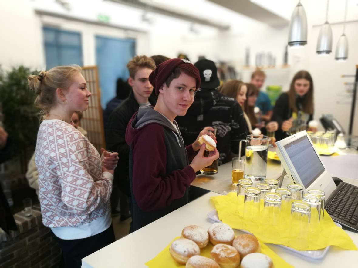 zu sehen ist ein Schüler, der einen Krapfen in der Hand hält. Rundherum stehen Schülerinnen und Schüler.