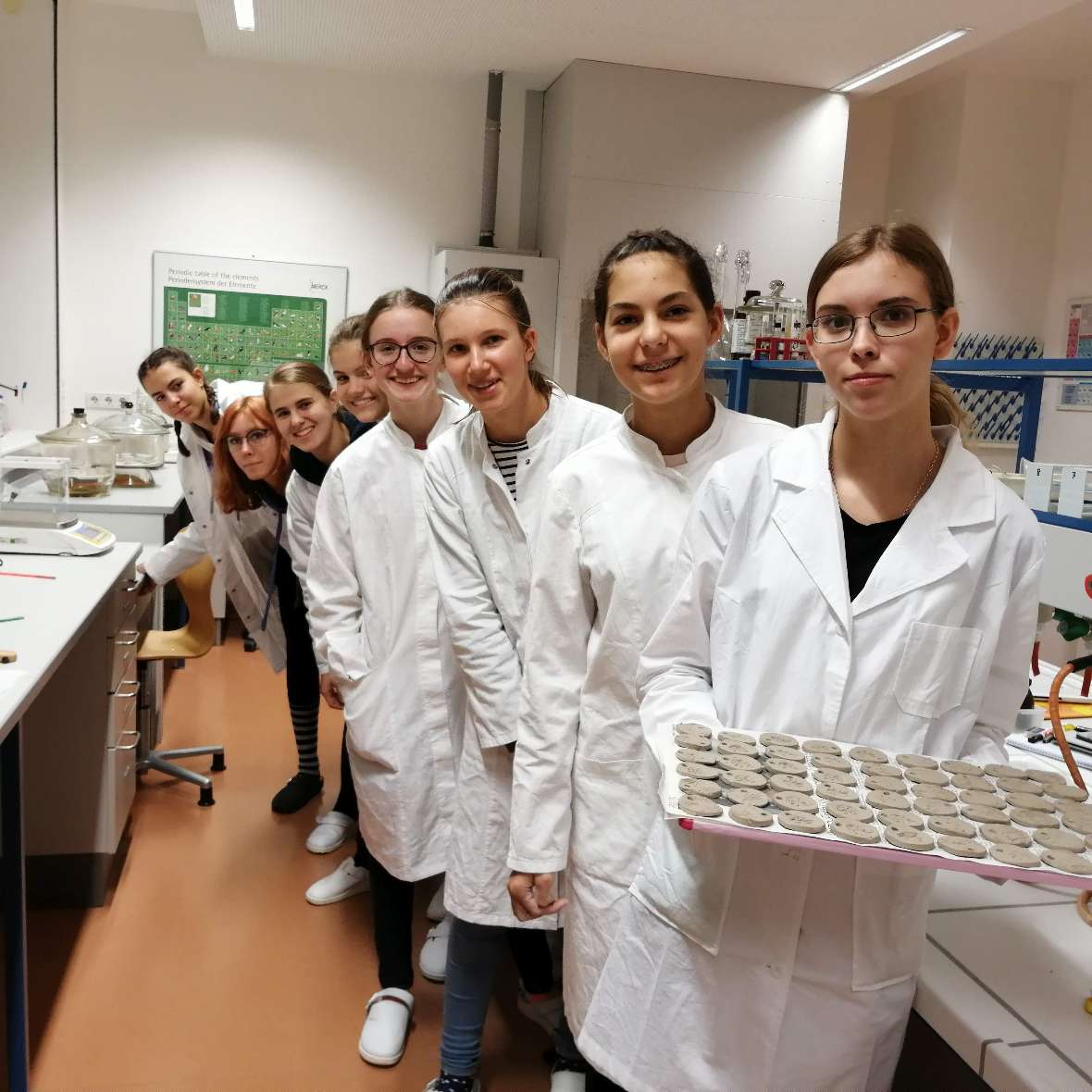 zu sehen sind Schülerinnen in weißen Arbeitsmänteln, die ein Tablett mit den rohen Ton-Medaillons in der Hand halten
