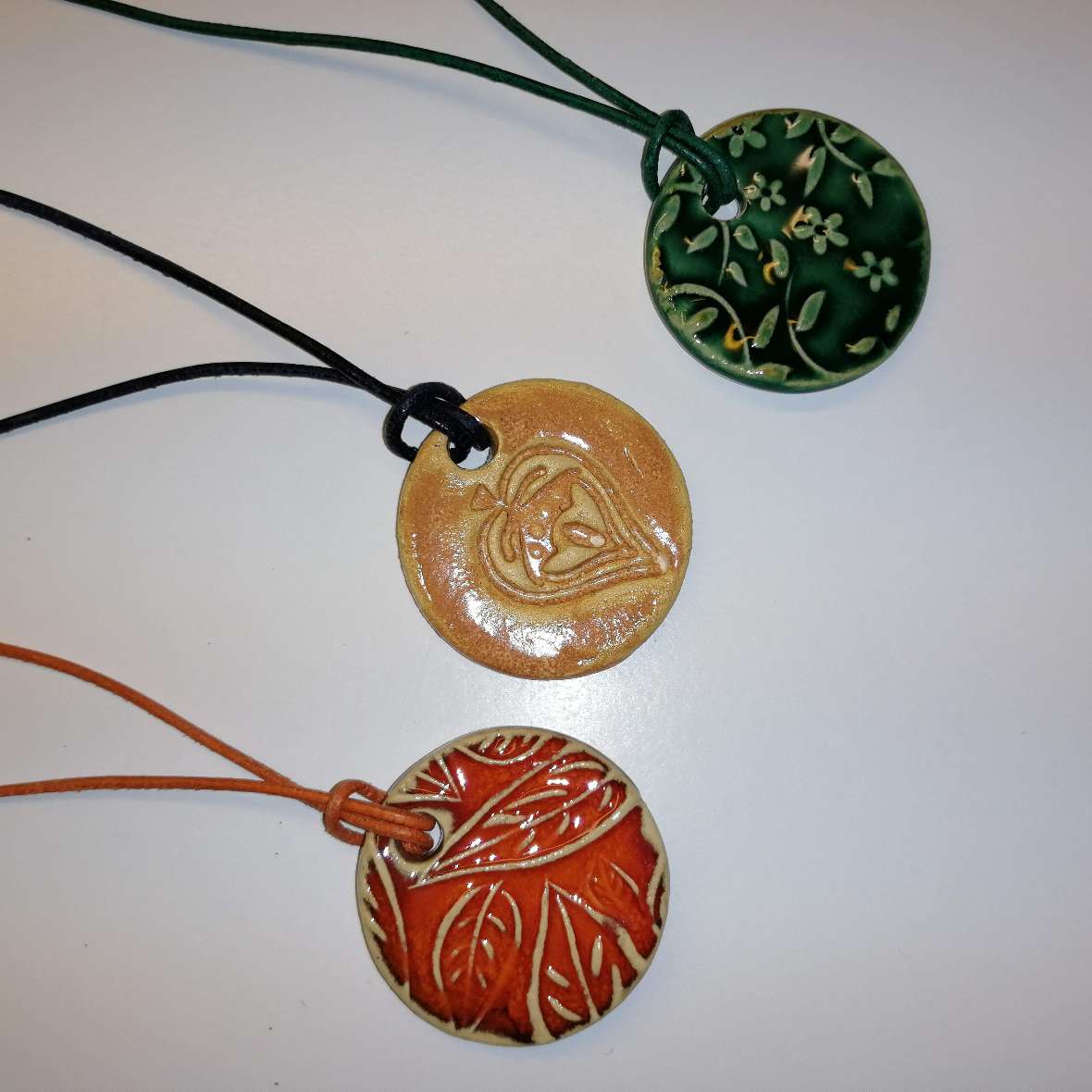 zu sehen sind 3 fertig gebrannte Blumen-Medaillons mit floralem Aufdruck in den Farben rot, gelb und grün. Die Medaillons sind an bunten Bändern befestigt.