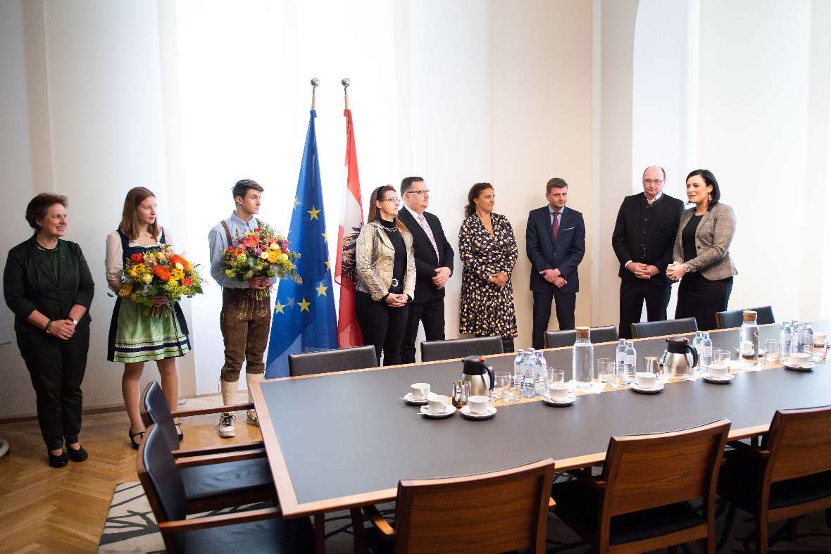 zu sehen sind Personen, die in einem großen Raum rund um einen sehr langen Tisch stehen. Ein Mädchen und ein Bursche haben einen Blumenstrauß in der Hand.