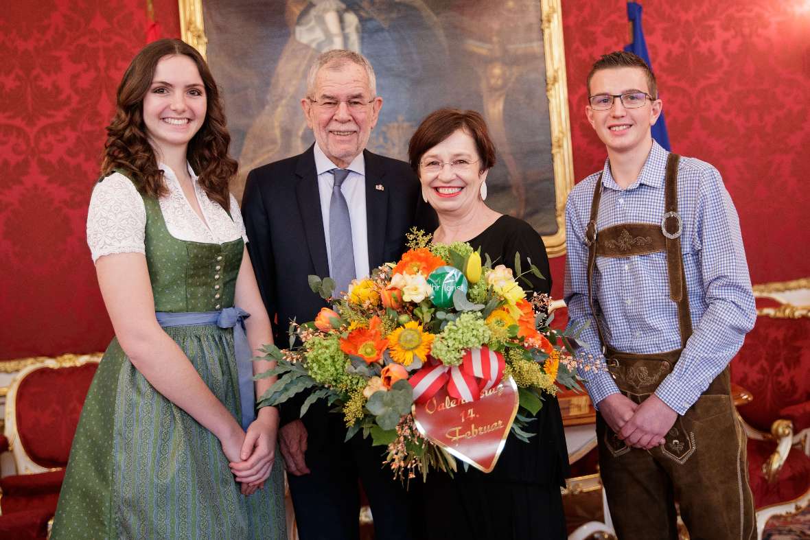 zu sehen in der Mitte des Bildes ist der Bundespräsident Van der Bellen mit seiner Frau, die einen bunten Blumenstrauß in der Hand hält; links und rechts daneben eine Schülerin und ein Schüler der Schule