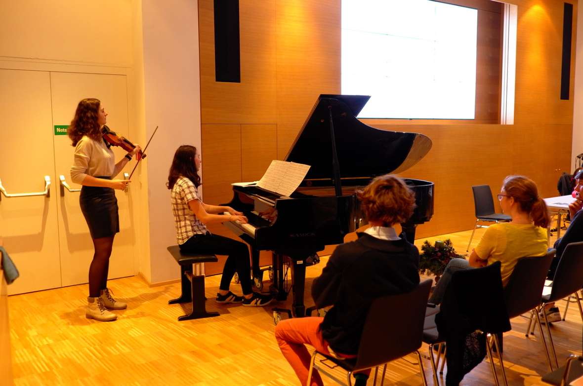 zu sehen ist ein Mädchen, das Klavier spielt, dahinter steht ein anderes Mädchen, das auf einer Geige spielt