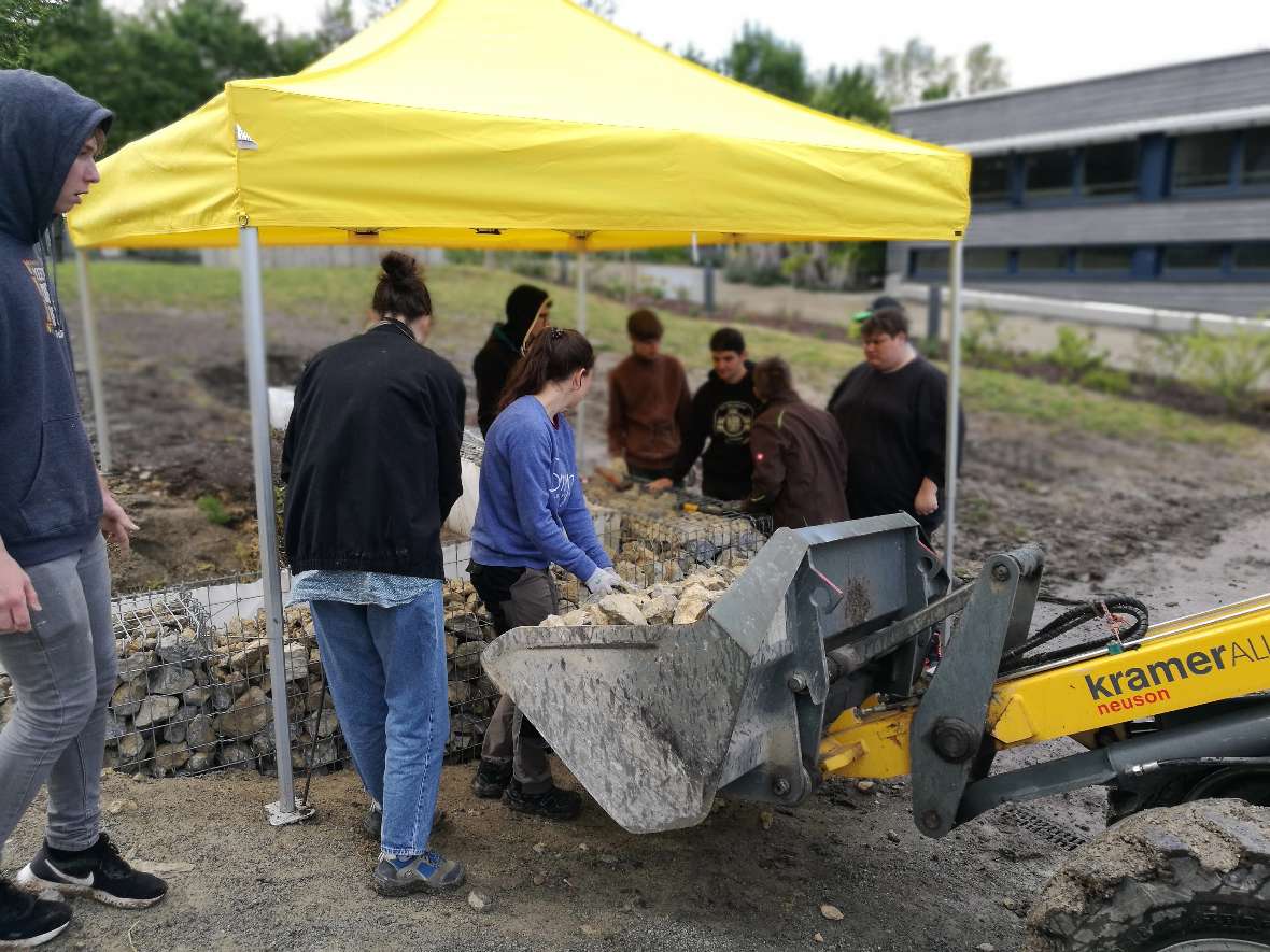 Schüler*innen befüllen die Drahtkörbe mit Grobschlag. Im Vordergrund steht ein Bagger.