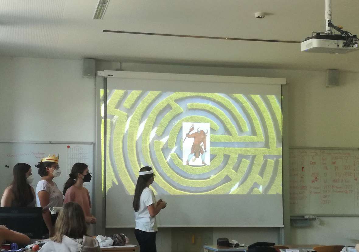Schülerinnen und Schüler halten ein Referat und stehen vor einer Beamerwand mit einer Abbildung eines Labyrinths und dem Minotaurus