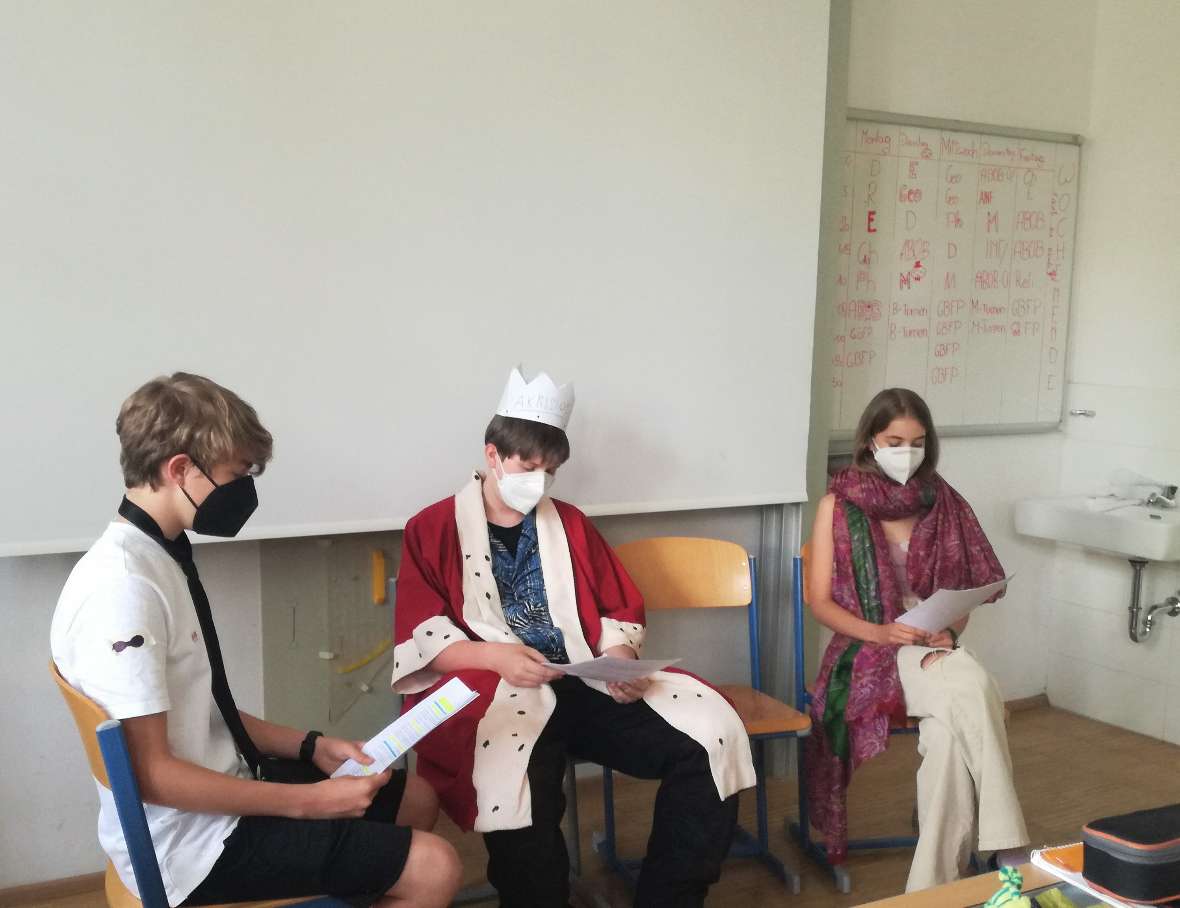 Zwei Schüler und eine Schülerin sitzen vor der Klasse, wobei der mittlere Schüler als König Akrisios verkleidet ist