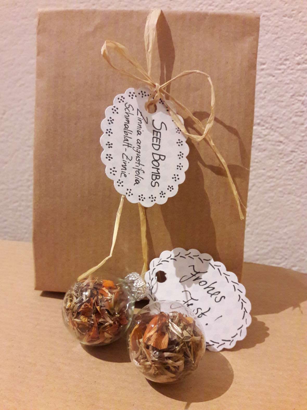 Seed-Bombs im selbstgemachten Papiersäckchen mit Geschenkanhänger und die fertigen Christbaumkugeln mit Samen und orangen Blütenblättern von Zinnia angustifolia