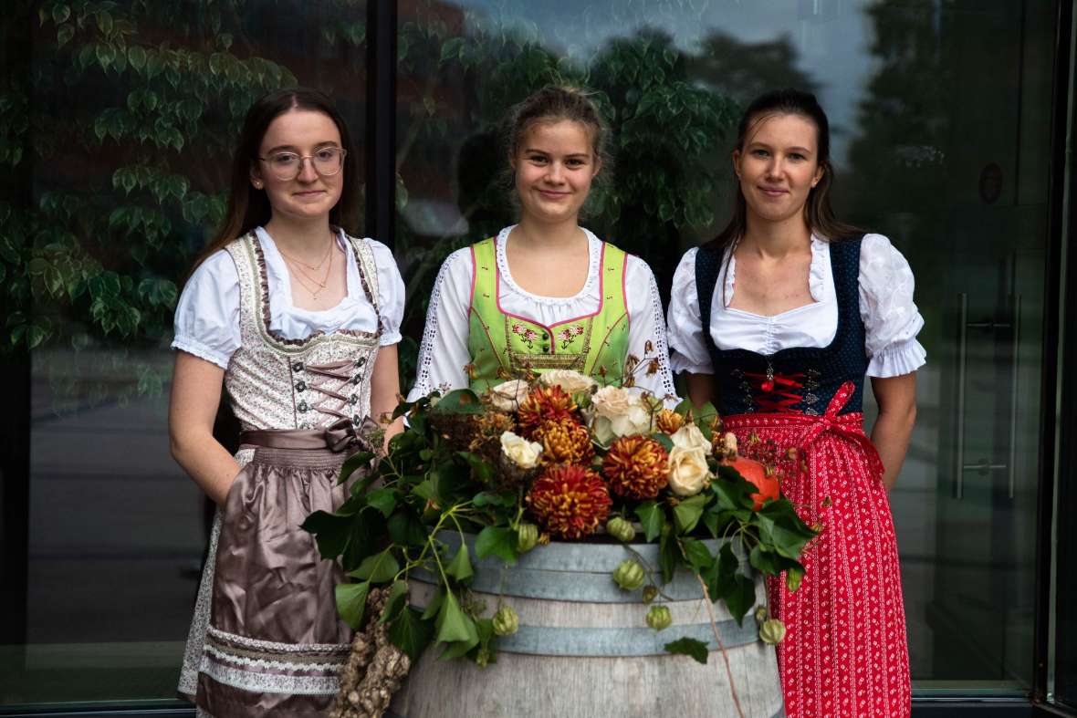 Drei Schülerinnen im Dirndl stehen vor einem Weinfass mit Blumendekoration