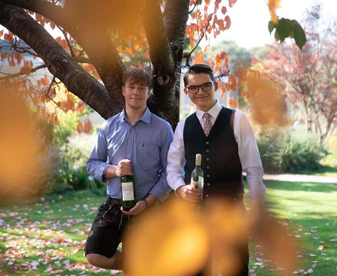 Zwei Schüler in Tracht stehen unter einem Baum und halten Weinflaschen in den Händen