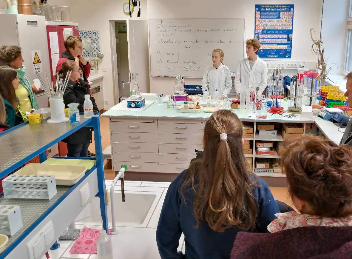 Eine Schülerin und ein Schüler im weißen Labormantel stehen im Labor und sind von Interessent:innen umgeben