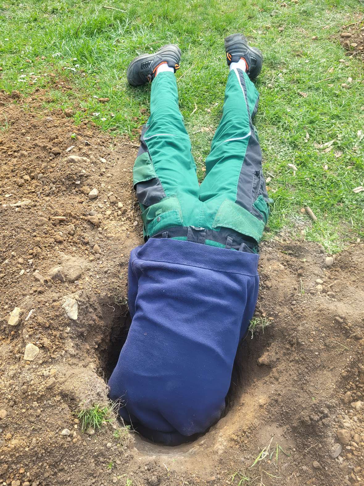 Schüler beim händischen Bodenaushub. Schüler liegt am Boden, Kopf und Schultern sind im Aushubloch verschwunden.