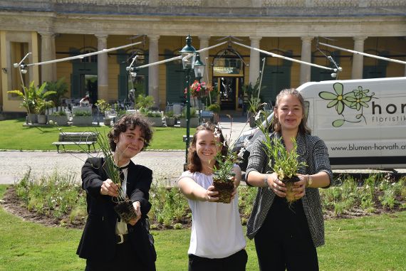Drei Schülerinnen halten jeweils eine Pflanze in der Hand und posieren vor einem Gebäude mit Säulen
