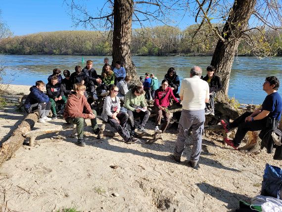 Schülerinnen und Schüler verweilen auf Baumstamm am Donauufer. Im Hintergrund die blaue Donau.
